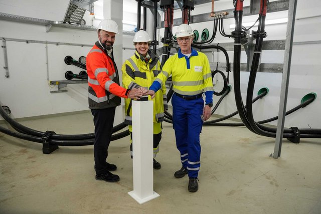 Drei Personen mit Schutzhelmen und Warnkleidung aktivieren gemeinsam per Knopfdruck eine technische Anlage in einem Umspannraum. Im Hintergrund verlaufen mehrere Hochspannungskabel und Anschlüsse an der Wand.