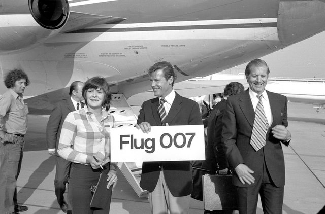 Roger Moore hält vor einem Flugzeug ein Schild mit der Aufschrift Flug 007 hoch.
