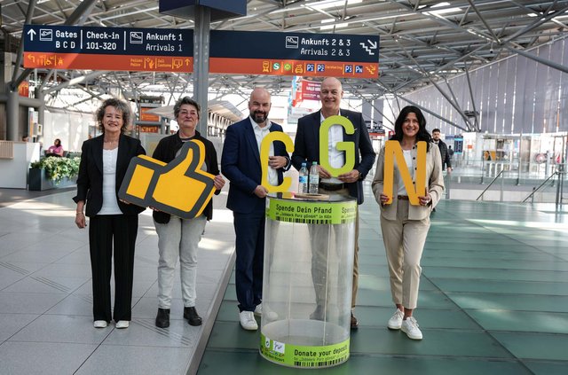 Fünf Vertreter von Köln Bonn Airport, JobWerk Porz und Grüner Punkt stehen im Terminal 2 des Köln Bonn Airport hinter einem grünen Pfandspendebehälter und feiern 10 Jahre Pfandprojekt 'Spende dein Pfand' am Flughafen Köln/Bonn.