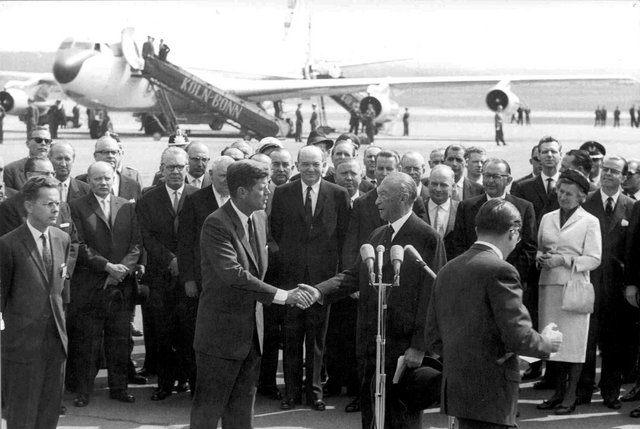 Konrad Adenauer schüttelt John F. Kennedy vor Zuschauern die Hand. Im Hintergrund ist ein Flugzeug zu sehen.