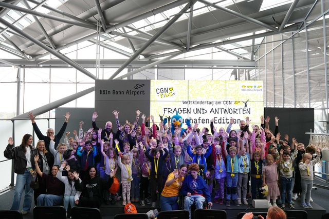 90 Kinder posieren winkend vor einer Präsentationswand des Flughafens
